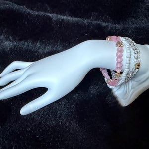 Puede incluir: Una colección de pulseras en tonos rosa, blanco y dorado, exhibidas en una mano de maniquí blanca. Las pulseras presentan cuentas, cristales y dijes en forma de corazón. El fondo es una superficie oscura y texturizada.