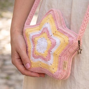 Op de afbeelding: Een roze, gele en witte gehaakte ster-vormige tas met een bijpassende riem. De tas heeft een ritssluiting en een kleine sterrenbedel. De tas wordt door een persoon vastgehouden.
