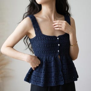 May include: A dark navy blue sleeveless crochet top featuring a square neckline, wide shoulder straps, and a peplum-style hem. The front of the garment is accented with three small, round white pearl-like buttons aligned vertically along the center.