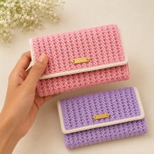 Puede incluir: Dos carteras rectangulares de ganchillo, una rosa y otra morada, cada una con un patrón de punto texturizado, un borde color crema y una pequeña placa de metal rectangular de tono dorado centrada en la solapa.
