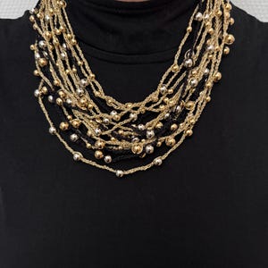 May include: A multi-strand necklace featuring gold, silver, and black beaded strands. The necklace has a layered design with small, round beads and a woven texture. The necklace is worn over a black top.