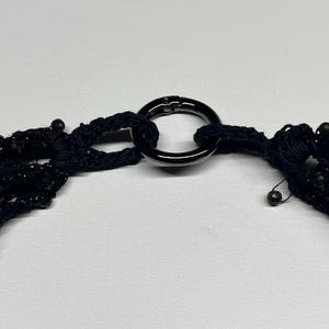 May include: A black necklace with multiple strands of beads and a large, dark metal ring clasp. The necklace is made of a woven material and has a delicate, intricate design. The clasp is a simple, yet elegant, circular shape.