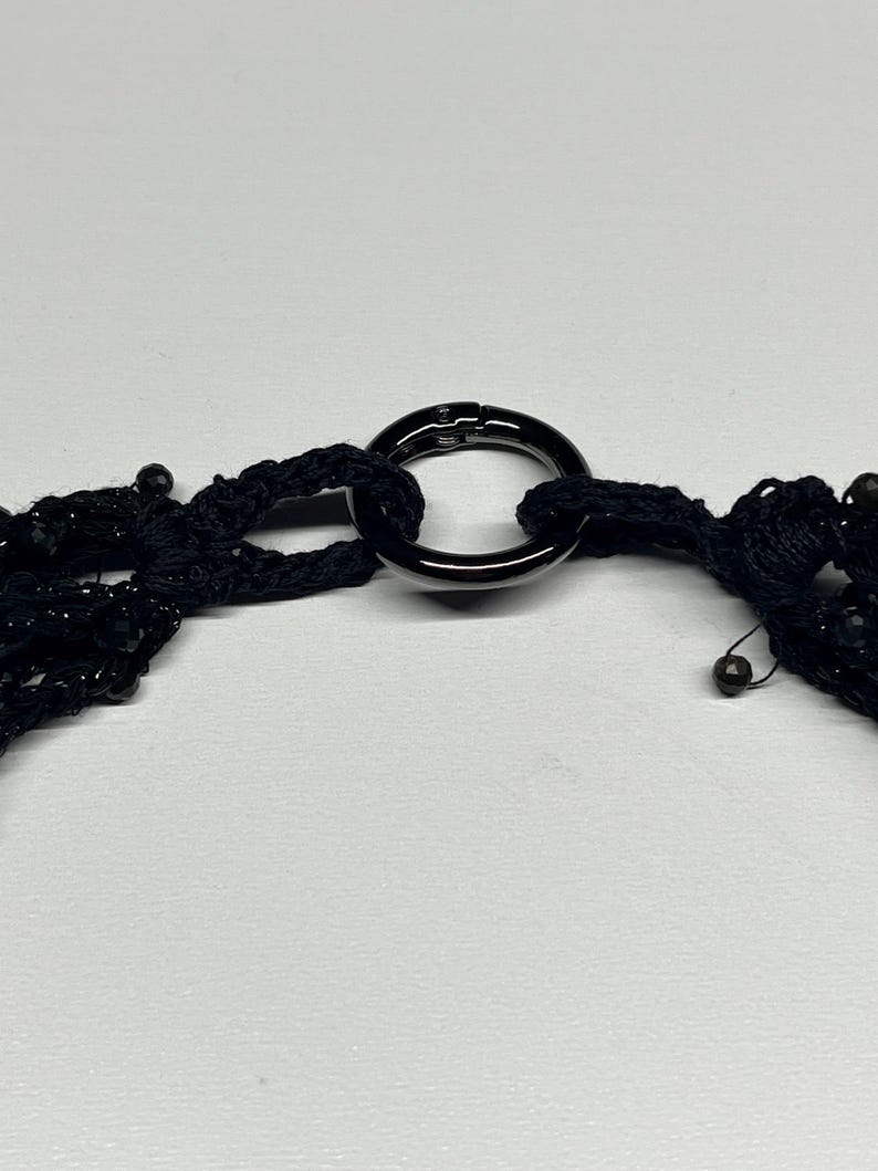 May include: A black necklace with multiple strands of beads and a large, dark metal ring clasp. The necklace is made of a woven material and has a delicate, intricate design. The clasp is a simple, yet elegant, circular shape.