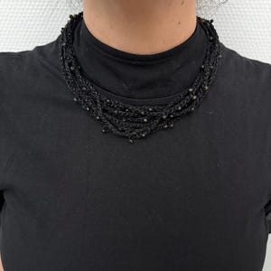 Könnte beinhalten: Eine mehrsträngige schwarze Halskette mit kleinen, dunklen Perlen. Die Halskette besteht aus gewebtem Material und liegt eng am Hals an. Die Person auf dem Bild trägt einen schwarzen Rollkragenpullover.