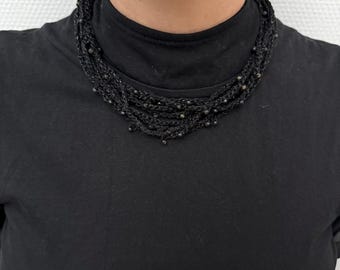 May include: A multi-strand black necklace with small, dark beads. The necklace is made of woven material and sits close to the neck. The person in the image is wearing a black turtleneck.