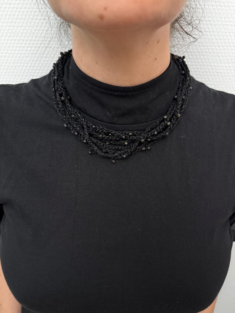 May include: A multi-strand black necklace with small, dark beads. The necklace is made of woven material and sits close to the neck. The person in the image is wearing a black turtleneck.
