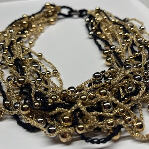 May include: A multi-strand necklace featuring alternating strands of gold and black. The necklace is adorned with small, shiny gold beads. The design is intricate and layered, creating a bold and elegant statement piece. The necklace is likely made of woven threads.