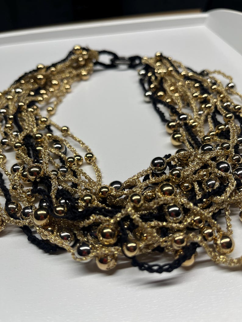 May include: A multi-strand necklace featuring alternating strands of gold and black. The necklace is adorned with small, shiny gold beads. The design is intricate and layered, creating a bold and elegant statement piece. The necklace is likely made of woven threads.