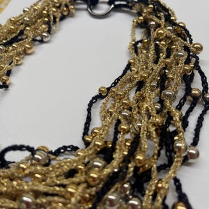 May include: A multi-strand necklace featuring gold and silver beads, interwoven with black and gold threads. The necklace has a black cord and a silver clasp. The beads are various sizes and the necklace is on a white background.