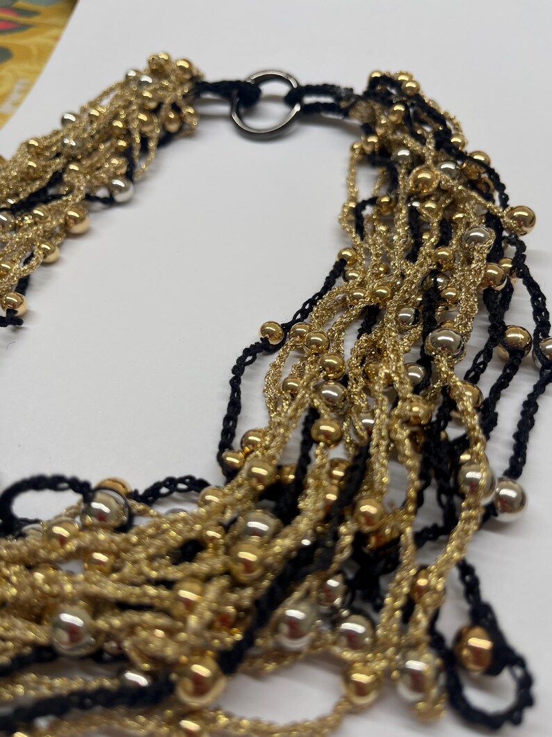 May include: A multi-strand necklace featuring gold and silver beads, interwoven with black and gold threads. The necklace has a black cord and a silver clasp. The beads are various sizes and the necklace is on a white background.