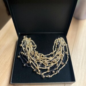 May include: A multi-strand necklace with gold-colored beads and dark blue strands, presented in a black box. The necklace features multiple layers of beaded strands, creating a layered effect. The beads are evenly spaced along the strands.