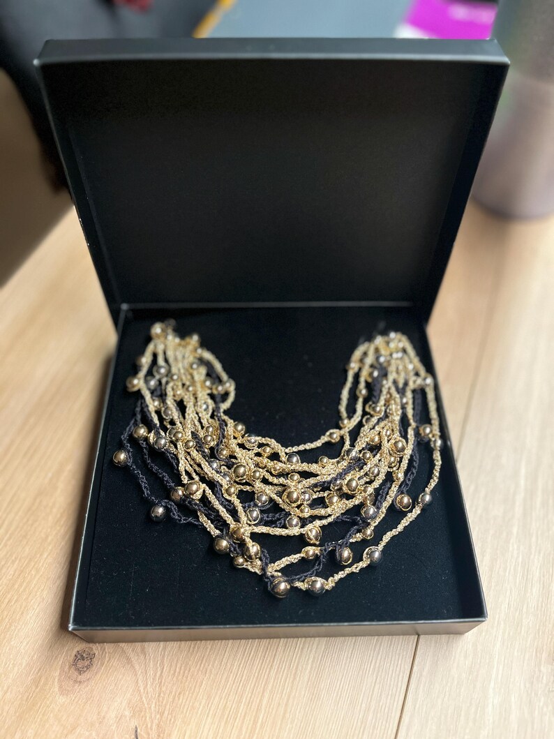 May include: A multi-strand necklace with gold-colored beads and dark blue strands, presented in a black box. The necklace features multiple layers of beaded strands, creating a layered effect. The beads are evenly spaced along the strands.
