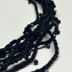 May include: Close-up of a black beaded necklace with multiple strands. The necklace features a woven design with small, dark beads interspersed throughout. The strands are twisted and hang loosely, creating a textured appearance. The background is plain white.