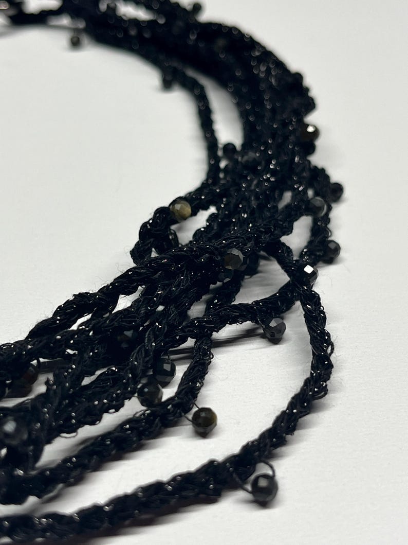 May include: Close-up of a black beaded necklace with multiple strands. The necklace features a woven design with small, dark beads interspersed throughout. The strands are twisted and hang loosely, creating a textured appearance. The background is plain white.