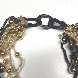 May include: Close-up of a necklace featuring strands of gold-colored beads and black woven threads. The necklace is connected by a dark metal ring clasp. The beads are various sizes and the overall design is intricate.