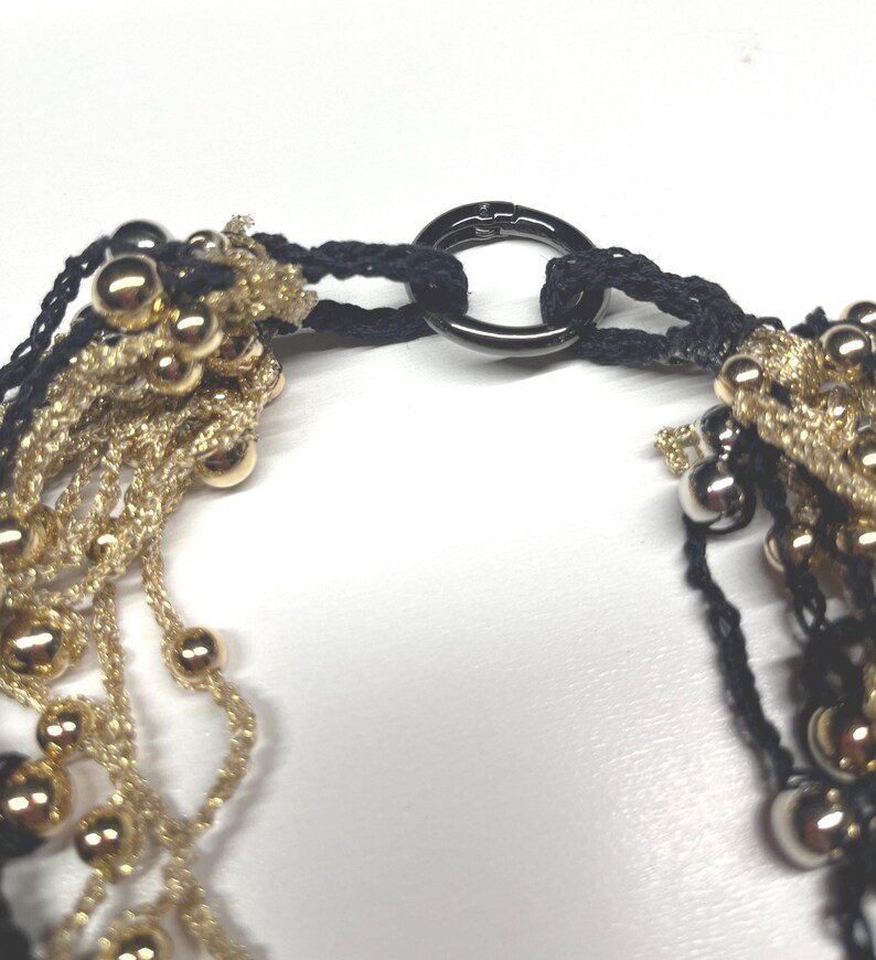 May include: Close-up of a necklace featuring strands of gold-colored beads and black woven threads. The necklace is connected by a dark metal ring clasp. The beads are various sizes and the overall design is intricate.