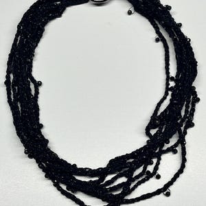 May include: Black beaded necklace with multiple strands. The necklace features a large, dark metal ring clasp. The strands are made of black thread and small, round beads.
