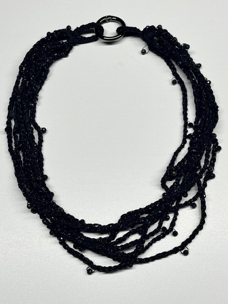 May include: Black beaded necklace with multiple strands. The necklace features a large, dark metal ring clasp. The strands are made of black thread and small, round beads.