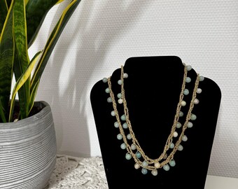 May include: A layered necklace with small, light blue and white beads. The necklace is displayed on a black velvet bust. The background includes a potted plant with green and yellow leaves and a white wall.