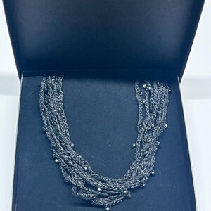 May include: A dark gray multi-strand necklace with small, silver-colored beads is displayed inside a black box. The necklace is made of woven strands and has a layered design. The box is open, revealing the jewelry.