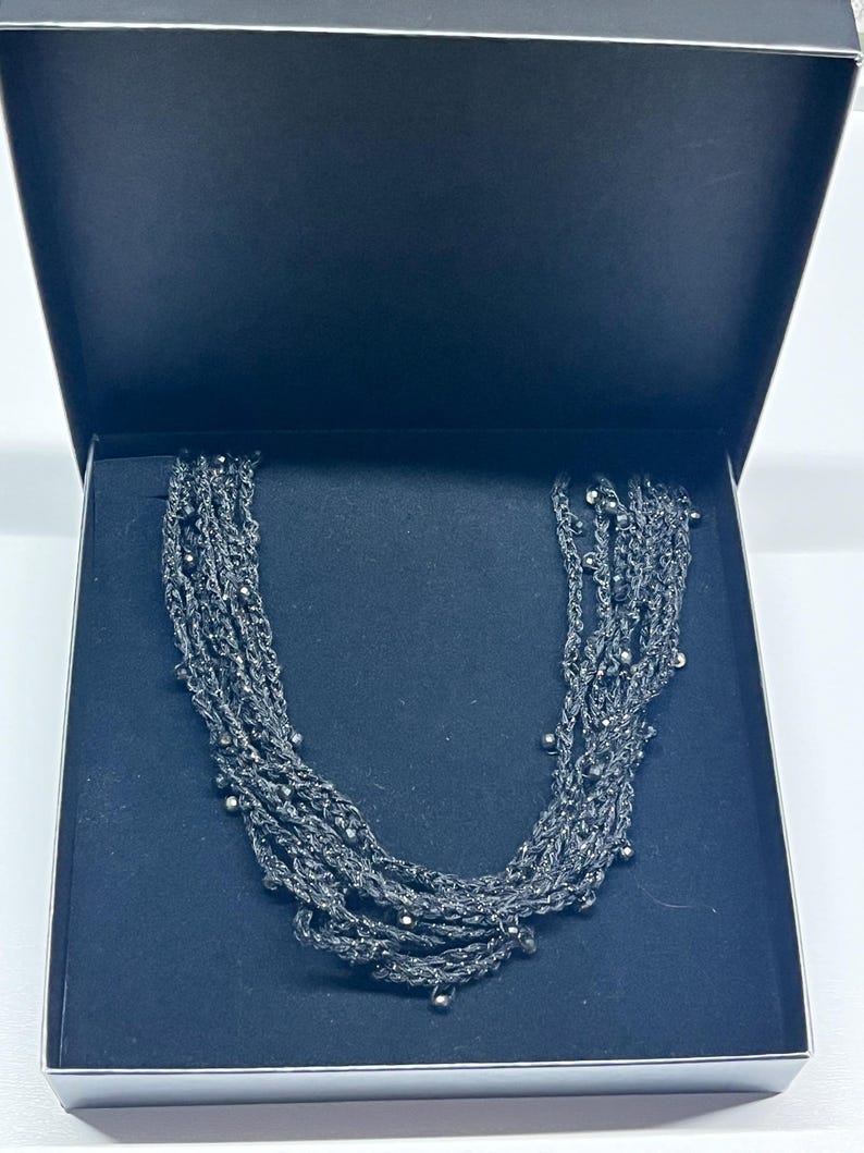 May include: A dark gray multi-strand necklace with small, silver-colored beads is displayed inside a black box. The necklace is made of woven strands and has a layered design. The box is open, revealing the jewelry.