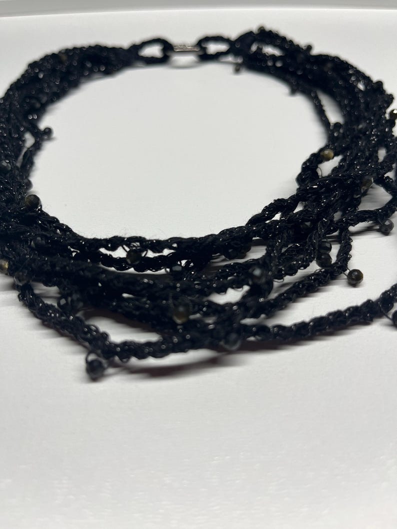 May include: Black multi-strand necklace with small beads. The necklace is made of a woven material and has a silver clasp. The necklace is a choker style and is suitable for formal or casual wear.
