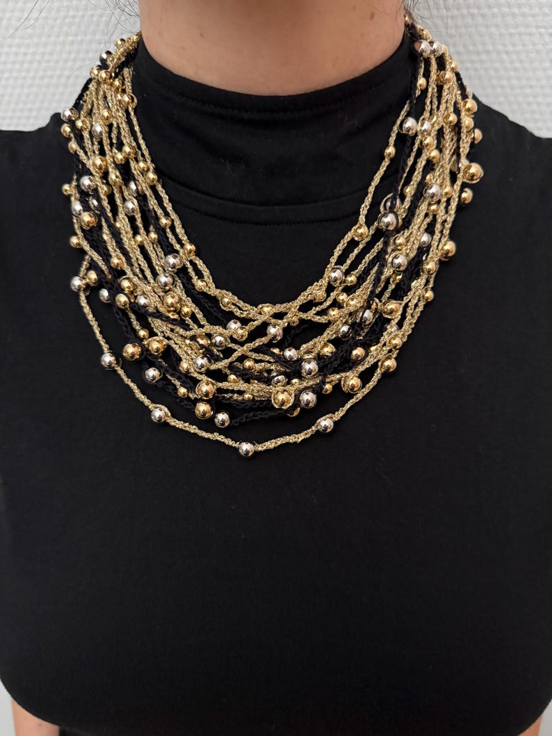 May include: A multi-strand necklace featuring gold, silver, and black beaded strands. The necklace has a layered design with small, round beads and a woven texture. The necklace is worn over a black top.