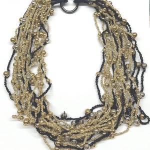 May include: A multi-strand necklace featuring gold, silver, and black beads. The necklace has a black cord and a silver-colored ring clasp. The strands are layered, creating a textured and elegant look. This statement necklace is suitable for various occasions.