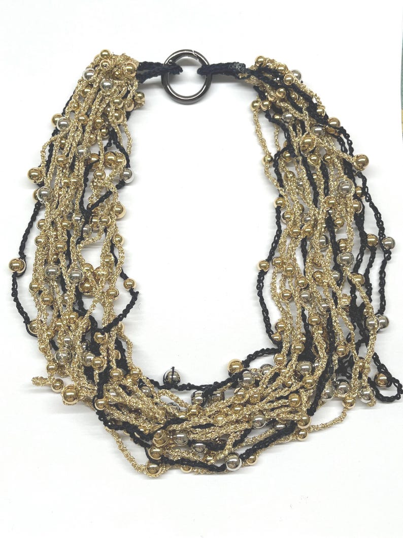 May include: A multi-strand necklace featuring gold, silver, and black beads. The necklace has a black cord and a silver-colored ring clasp. The strands are layered, creating a textured and elegant look. This statement necklace is suitable for various occasions.