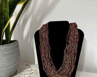 May include: A multi-strand necklace displayed on a black stand. The necklace features strands of burgundy and gold-colored beads, with small red beads interspersed throughout. The necklace is a statement piece.