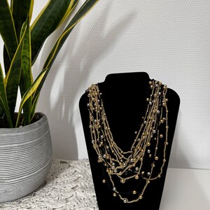 May include: A multi-strand necklace displayed on a black jewelry bust. The necklace features strands of gold and silver beads, creating a layered effect. A potted plant with green and yellow leaves is in the background.