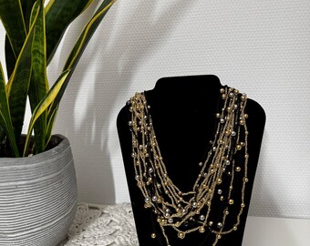 May include: A multi-strand necklace displayed on a black jewelry bust. The necklace features strands of gold and silver beads, creating a layered effect. A potted plant with green and yellow leaves is in the background.