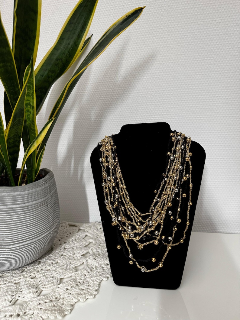 May include: A multi-strand necklace displayed on a black jewelry bust. The necklace features strands of gold and silver beads, creating a layered effect. A potted plant with green and yellow leaves is in the background.