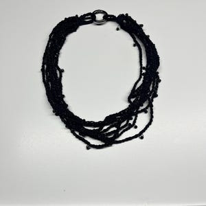 May include: A black multi-strand necklace with a silver-tone clasp. The necklace is made of multiple strands of black material, possibly thread or cord, and features small, dark beads. The clasp is a simple, circular design.