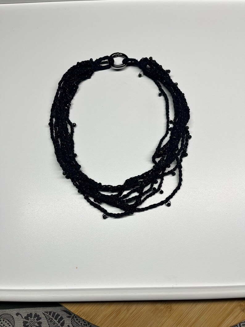 May include: A black multi-strand necklace with a silver-tone clasp. The necklace is made of multiple strands of black material, possibly thread or cord, and features small, dark beads. The clasp is a simple, circular design.