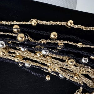 May include: Close-up of layered necklaces featuring gold and silver metallic beads on black and gold cords. The beads are evenly spaced along the cords, creating a textured and elegant look. The necklaces are displayed against a black background.