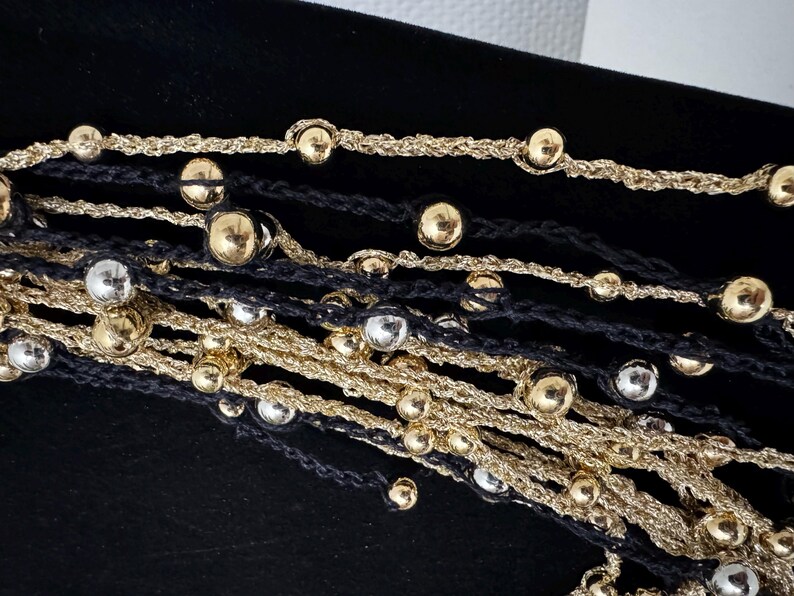 May include: Close-up of layered necklaces featuring gold and silver metallic beads on black and gold cords. The beads are evenly spaced along the cords, creating a textured and elegant look. The necklaces are displayed against a black background.
