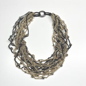 May include: A multi-strand necklace featuring alternating strands of black and gold-colored beads. The necklace has a circular clasp and is set against a white background. The beads are small and evenly spaced, creating a textured appearance.