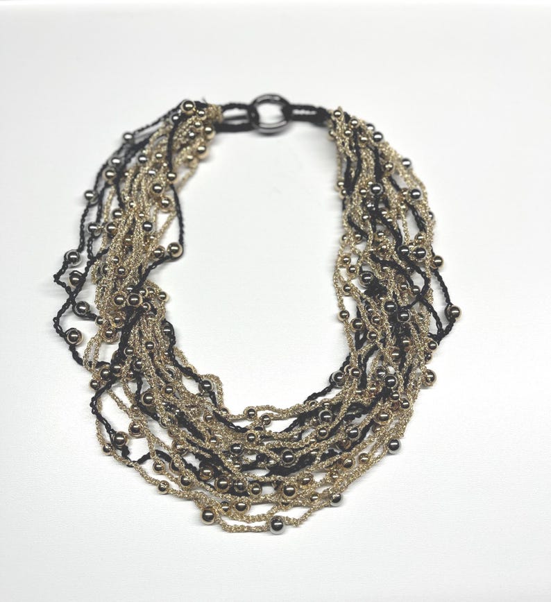 May include: A multi-strand necklace featuring alternating strands of black and gold-colored beads. The necklace has a circular clasp and is set against a white background. The beads are small and evenly spaced, creating a textured appearance.