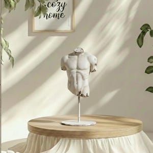 Könnte beinhalten: Eine weiße, verwitterte Torso-Statue auf einem Metallständer. Die Skulptur wird auf einem runden Holztisch mit einem weißen Stoffrock ausgestellt. Der Hintergrund enthält ein gerahmtes Schild mit der Aufschrift "cozy home".