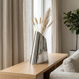 May include: A modern, silver-colored vase with a unique, angled design. The vase holds dried wheat and pampas grass, adding a natural element. The vase sits on a light-colored wooden table, creating a minimalist aesthetic.