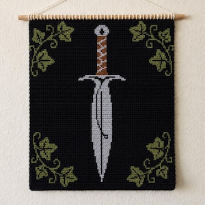 May include: A rectangular crochet wall hanging featuring a black background with a central silver dagger design. The dagger has a brown handle with a white zigzag pattern. Four green ivy leaf clusters are positioned in each corner of the piece.