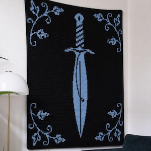 May include: A rectangular black wall hanging featuring a large, centred light blue sword graphic. The sword is flanked by four symmetrical light blue vine patterns in each corner, all created with a pixelated, grid-like texture design.
