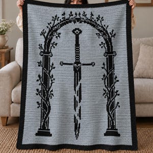 May include: A rectangular crochet blanket featuring a light gray background with a black border. The center design displays a black graphic of a sword positioned vertically inside a tall, arched stone-like gateway entwined with leafy vines.