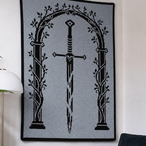 May include: A rectangular crochet wall hanging featuring a light grey background with a black border. The design depicts a central broken sword positioned beneath a tall, ornate archway adorned with climbing vines and leaves in black yarn.