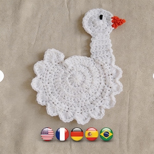 May include: A handmade white crochet goose-shaped coaster with a red and black beak. The coaster has a circular body with a scalloped edge and a long neck. The coaster is on a beige surface.