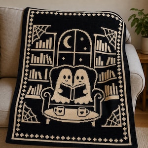 May include: A black and cream crochet blanket featuring a central design of two ghosts sitting in an armchair reading a book. The scene includes bookshelves, a window with a crescent moon, spiderwebs in the corners, and a table with two mugs.