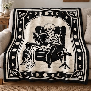May include: A black and white crochet blanket featuring a central graphic of a skeleton sitting in an armchair reading a book. The border is decorated with a repeating moon phase pattern and geometric corner accents, creating a high-contrast design.