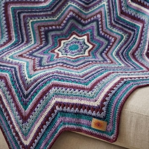 May include: A handmade, star-shaped crochet blanket in shades of teal, purple, and white. The blanket features a detailed, layered design with a central star motif and a decorative border. The blanket is resting on a neutral-coloured surface.