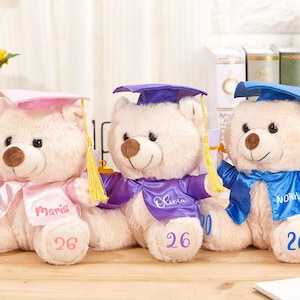 Könnte beinhalten: Drei Plüsch-Abschlussbären. Jeder Bär trägt eine Abschlusskappe und einen Umhang in Rosa, Lila und Blau. Die Bären haben Diplome und die Namen "Maria", "Olivia" und "Noah" sind auf ihren Umhängen aufgedruckt. Die Zahl "26" ist auf die Füße der Bären gedruckt.